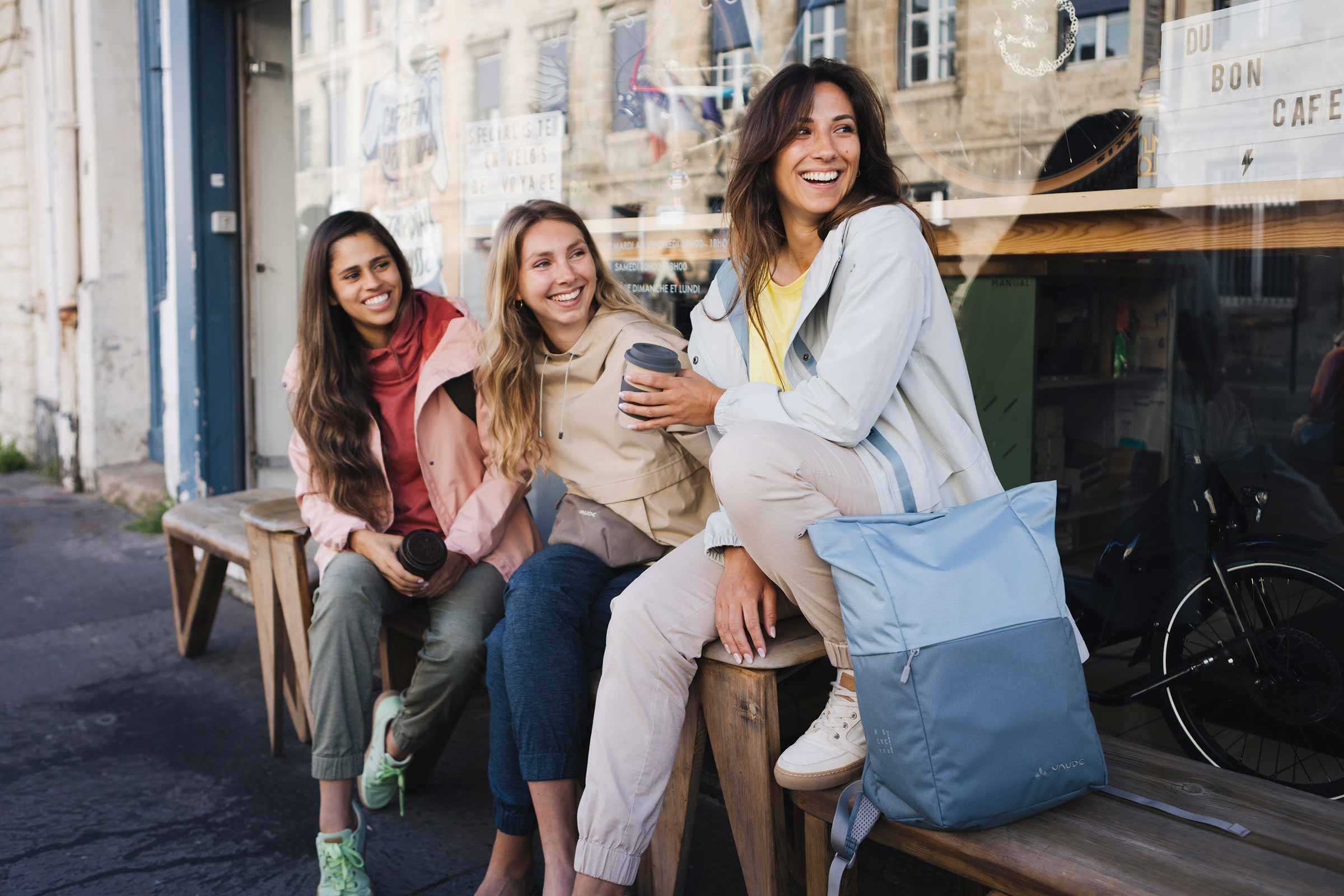 three women with bags and backpacks sitting in front of a store